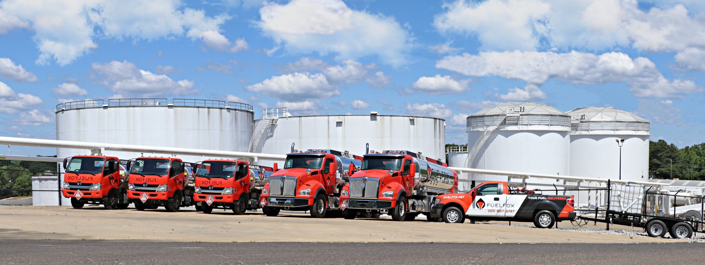 The FuelFox fleet lined up at a fuel terminal: three box trucks, two tankers and a service pickup, with storage tanks behind them.